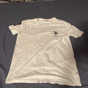 Vit t-shirt från U.S. Polo Assn. - Vit t-shirt från U.S. Polo Assn. med klassisk rund hals och korta ärmar. På bröstet finns en snygg broderad logga med poloryttare och texten USPA. Materialet är mjuk bomull och nederkanten har en liten detalj med färgband. Perfekt för en chill och stilren look.