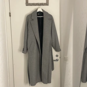 Grå lång kappa från Zara, stl S - Snygg grå kappa från Zara i klassisk modell med bred krage och knytskärp i midjan. Lång passform, två fickor framtill och knappar vid ärmslut. Perfekt för dig som gillar stilrena och tidlösa plagg. (Aldrig använd)