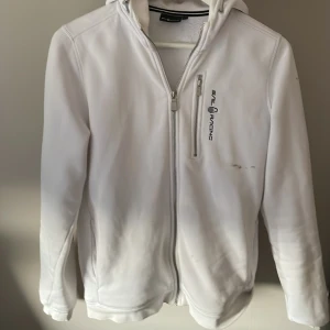 Sail Racing hoodie - Snygg vit hoodie från Sail Racing med huva och dragkedja framtill. Jackan har en bröstficka med dragkedja och logga på bröstet. Perfekt för blåsiga dagar och enkel att matcha med det mesta. Tröjan har olje fläckar på få ställen som på första bilden därav pris. 176 men passar M