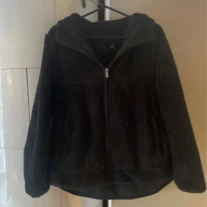 Svart teddykofta med dragkedja - Mysig svart teddykofta med hel dragkedja framtill och två fickor. Jackan har en fluffig och mjuk känsla, perfekt för kyliga dagar. Långärmad och med en avslappnad passform som är enkel att slänga på sig över en hoodie eller t-shirt.