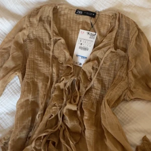 Helt ny Beige Blus med Volang från Zara - Supersnygg beige blus från Zara i storlek S, med prislapp (original pris är 380kr). Blusen har fina volang detaljer framtill och på ärmsluten, samt snygg snörning vid halsen. Materialet är lätt och luftigt. Ärmarna är långa med volangkant och broderade detaljer. Säljer då den aldrig har kommit till användning 🩷🩷