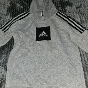 Grå Adidas hoodie med svarta ränder - Säljer en grå hoodie från Adidas med klassiska svarta ränder längs axlarna och ett stort svart Adidas-tryck på bröstet. Tröjan har huva, känguruficka och är långärmad. Perfekt för chill eller träning, riktigt skön och stilren.