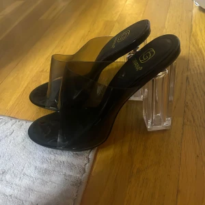 Svarta klackskor från Givana med transparent klack - Snygga svarta klackskor från Givana med bred rem i transparent plast och öppen tå. Skorna har en hög, fyrkantig klack i genomskinlig plast som ger en modern vibe. Perfekta för dig som vill sticka ut med en cool och trendig look.