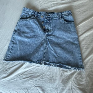 Blå jeanskjol från Pieces - Snygg blå jeanskjol från Pieces med rå kant nertill och knappar framtill. Kjolen har klassiska fickor både fram och bak samt hällor för bälte. Perfekt för dig som gillar en avslappnad och trendig stil. Mig waist