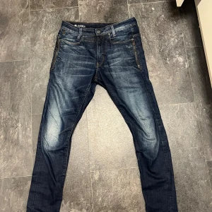 G-Star RAW D-Staq mörkblå jeans - Snygga mörkblå jeans från G-Star RAW, modell D-Staq med dragkedjedetaljer vid fickorna och coola slitningar. Jeansen har klassisk femficksdesign, kontrastsömmar och smal passform. Perfekta för dig som gillar streetwear och vill ha en modern look.