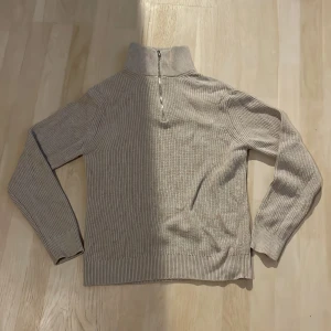 Beige ribbad stickad tröja half zip - Beige stickad tröja från EAST WEST i storlek S. Tröjan har ribbad struktur, hög krage och en snygg half zip-dragkedja framtill. Perfekt för lager-på-lager och chill höst- eller vinterstil.