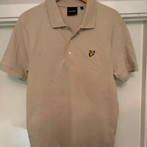 Beige pikétröja från Lyle & Scott - Snygg beige pikétröja från Lyle & Scott med klassisk krage och korta ärmar. Tröjan har tre knappar framtill och den ikoniska gula fågel-loggan broderad på bröstet. Perfekt för en clean och stilren look. Använd ett fåtal gånger. Skicket är 8/10