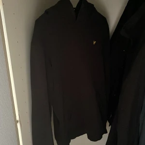 Svart hoodie från Lyle & Scott - Svart hoodie från Lyle & Scott med klassisk huva och lång ärm. Tröjan har en liten gul logga broderad på bröstet och är tillverkad i mjukt bomullsmaterial. Perfekt för en chill och stilren look.