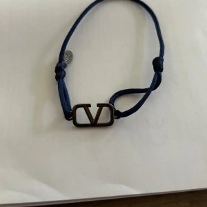 Valentino blå armband med V-logga - Snyggt armband från Valentino med mörkblå justerbar snodd och metallberlock med ikoniska V-loggan. Perfekt accessoar för dig som gillar stilrena detaljer och designer vibes. Metallbrickan har en diskret gravyr och armbandet är lätt att justera för bästa passform.