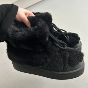 Svarta fluffiga boots med snörning H&M - Säljer ett par svarta boots med fluffig pälsimitation och snörning framtill. Skorna har en platt sula och är perfekta för kalla dagar när du vill vara både varm och snygg. De har en chunky look och känns riktigt mysiga.