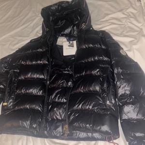 Svart Moncler jacka  - Säljer en svart, glansig dunjacka från Moncler med huva och dragkedjor. Jackan har klassisk Moncler-logga på ärmen, quiltad design och snygga detaljer som snörning vid huvan. Perfekt för kalla dagar och riktigt stilren look. Den är i S men massor M med 