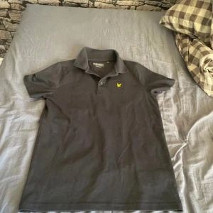 Svart pikétröja från Lyle & Scott - Svart pikétröja från Lyle & Scott med klassisk krage och knappslå framtill. Tröjan har ett gult broderat logomärke på bröstet och är tillverkad i en mjuk bomullsblandning med lite polyester och elastan för extra komfort. Perfekt för en chill och stilren look.