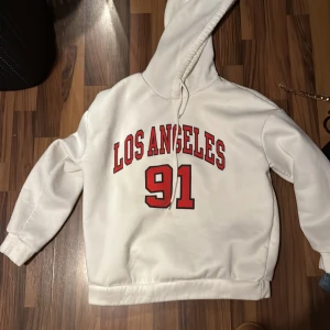Vit hoodie med Los Angeles-tryck - Säljer en vit hoodie med rött Los Angeles-tryck och siffran 91 på bröstet. Tröjan har huva med snörning och långa ärmar. Perfekt för dig som gillar streetstyle och sportig vibe. Passar chill dagar och när du vill ha en avslappnad look.