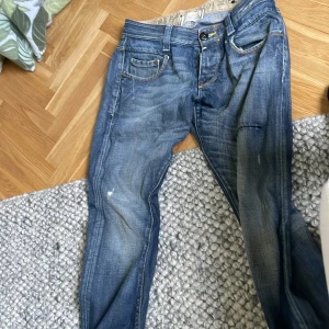 Blå jeans från G-Star Raw - Säljer ett par blå jeans från G-Star Raw med snyggt slitna detaljer och kontrastsömmar. Jeansen har klassisk femficksdesign, orange sömmar och unika broderier på bakfickorna. Perfekta för dig som gillar en avslappnad och cool stil.