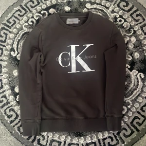  Calvin Klein Jeans - Snygg svart sweatshirt från Calvin Klein Jeans med stor vit logga framtill. Klassisk rund halsringning och långa ärmar. Perfekt för en clean och stilren look. Mjuk och skön bomullsblandning som passar året runt.