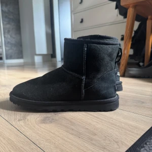 Svarta UGG boots i mocka - Mysiga svarta UGG boots i mjuk mocka med rund tå och platt sula. Klassisk modell med UGG-logga bak och kort skaft. Perfekta för kalla dagar och riktigt sköna att glida runt i hemma eller ute.