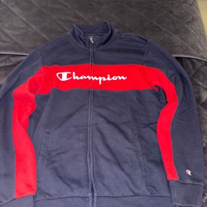 Champion ziptröja i marinblått och rött - Säljer ett snyggt sett med både ziptröja från Champion samt mjukisbyxor. Tröjan har hög krage, hel dragkedja och två fickor framtill. Perfekt för dig som gillar sportig stil. Mjukisbyxorna har fickor vid sidan och passar dig som är 13-14 år. 