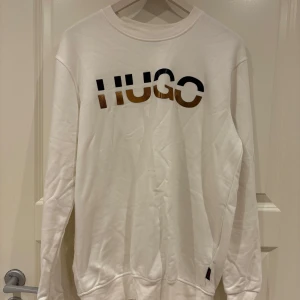 Vit sweatshirt från Hugo Boss - Snygg vit sweatshirt från Hugo Boss med stor logga i svart och guld framtill. Tröjan har rund halsringning och långa ärmar, perfekt för en clean och stilren look. Mjuk bomullskänsla och relaxed fit.