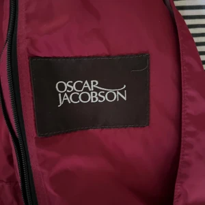 Vinröd vindjacka från Oscar Jacobson - Snygg vinröd vindjacka från Oscar Jacobson med huva och dragsko i midjan. Jackan har dragkedja framtill och är tillverkad i lätt polyester, perfekt för blåsiga dagar. Enkel och stilren design som passar till många outfits.