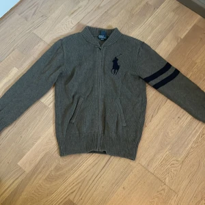 Grå kofta från Polo Ralph Lauren - Snygg grå kofta från Polo Ralph Lauren med dragkedja framtill och ribbade muddar. Jätte fin kvalité. 