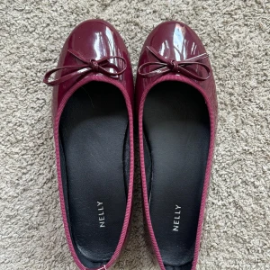 Vinröda ballerina från Nelly - Snygga vinröda ballerina från Nelly i glansigt syntetmaterial. Skorna har rund tå, platt sula och en söt rosett framtill. Perfekta för dig som vill ha en klassisk men trendig look med en touch av färg.