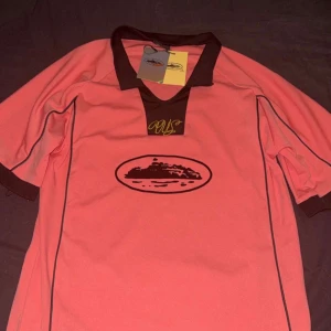 Corteiz talismo fotbollströja  - Snygg rosa fotbollströja från Artcutic i storlek small. Tröjan har svart krage och svarta detaljer längs ärmarna och sidorna. På bröstet finns ett svart tryck med motiv och en gul signatur. Materialet är lätt och andas, perfekt för träning eller match.