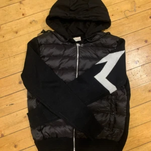 Moncler cardigan som ny väldigt fräsch  - Svart hoodie från Moncler med vadderad front och stickade ärmar. Stor vit blixtdetalj på ena ärmen och klassisk Moncler-logga. Dragkedja framtill och huva. Perfekt för dig som vill ha en streetig och exklusiv look.       Väldigt fräsch som ny storlek M. Väldigt sällsynt jacka och väldigt efterfrågad. Först till kvarn.