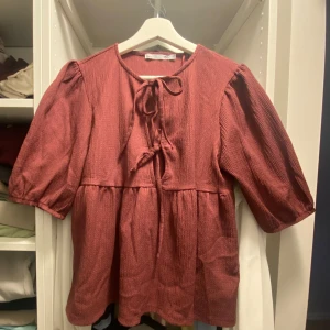 Roströd blus med knyt och puffärm - Supersöt roströd blus från lager 157 med puffiga halvlånga ärmar och knyt fram i halsen. Blusen har en lös passform och lätt rynkade detaljer under bysten för en flowy look. Perfekt till jeans eller kjol för en chill men ändå stilsäker vibe.