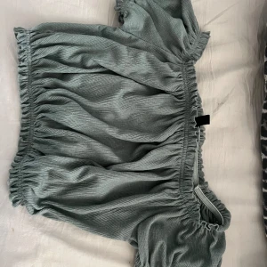 Grön offshoulder blus med volang - Supersöt grön blus med offshoulder och volangdetaljer. Toppen har en loose passform och är gjord i ett mjukt, ribbat material som ger en avslappnad vibe. Perfekt för dig som gillar trendiga och bekväma plagg med lite extra volym.