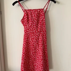 Röd blommig kortklänning med rosett - Supersöt röd kortklänning med vita blommor, smala axelband och en snygg rosettdetalj i ryggen. Klänningen har dragkedja bak för att ha det lättare att ta av och på och är perfekt för varma dagar. Materialet känns lätt och luftigt, och modellen är figurnära upptill med utsvängd kjol. Den är använd 1-2 gånger