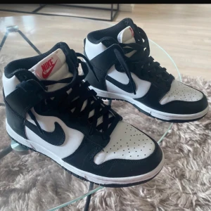 Nike Dunk panda sneakers - Nike Dunk High sneakers i svart och vitt med klassisk röd Nike-logga på plösen. Skorna har snörning, rund tå och platt sula. Ovandelen är i skinn, och den höga siluetten ger en sportig vibe. Perfekta för dig som gillar streetstyle och ikoniska sneakers. Använd 1 gång, mycket fint skick