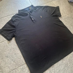 Ralph Lauren pike - Svart pikétröja från Polo Ralph Lauren i slim fit-modell. Klassisk krage, tre knappar och broderad logga på bröstet. Tillverkad i mjuk bomull, perfekt för en clean och stilren look. Kortärmad och passar dig som gillar tidlös stil. Strl XXL men sitter snarare som XL/L