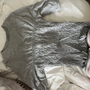 Grå ribbad topp med smockdetalj - Säljer en grå topp med ribbad struktur upptill och smockad midja. Toppen har rund halsringning och korta ärmar. Perfekt för dig som gillar en enkel men ändå unik stil. Säljer även i svart o vit oxå