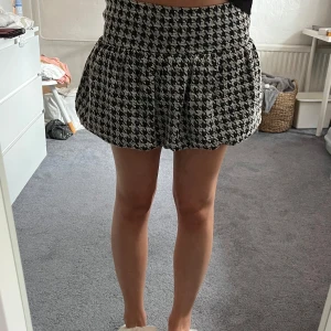 Svartvit rutig kortkjol från Zara - Supersöt kortkjol från Zara i svartvitt rutigt mönster. Kjolen har är ballongformad siluett och är gjord i ett grovt, stickat material som ger en cool och trendig vibe. Perfekt att styla med en oversized tröja eller boots för en snygg look. Köpt för 299kr. 