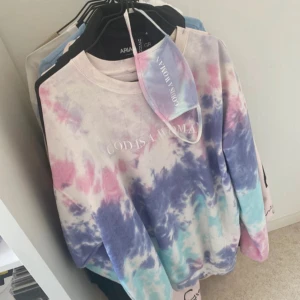 God is a woman crewneck - Superfin oversized sweatshirt i pastell tie-dye med texten 'God is a Woman' framtill och ansiktsmask. Tröjan har långa ärmar och en relaxed fit, perfekt för dig som gillar färg och statement-plagg. Kommer från Ariana Grandes merch.