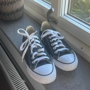 Svarta Converse All Star sneakers - Klassiska svarta Converse All Star sneakers med vit sula och vita snören. Lågt skaft, rund tå och tydliga kontrastsömmar. Ovandel i textil och sula i gummi. Perfekta för en avslappnad och cool stil.