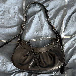 Svart axelväska i skinn  - Snygg svart axelväska i glansigt skinn med coola sömdetaljer och justerbar axelrem. Väskan har snörningsdetalj framtill och silvriga metalldetaljer som ger en trendig vibe. Perfekt storlek för det viktigaste och passar till många outfits.