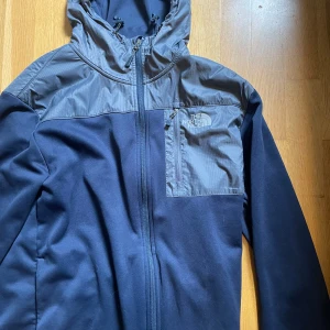 Blå hoodie från The North Face M - Snygg blå hoodie från The North Face med grå detaljer på axlar och huva. Hoodien har dragkedja framtill, bröstficka med dragkedja och justerbar huva. Perfekt för outdoor vibes.