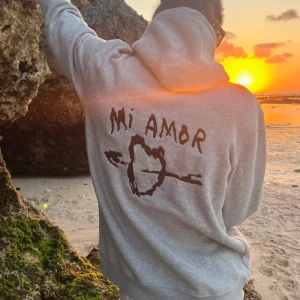 Grå hoodie Mira Paris med tryck - Grå hoodie från Mira Paris med svart tryck på ryggen där det står 'Mi Amor' och en pil genom ett hjärta. Tröjan har huva med snörning och en stor magficka. Mjuk och skön bomullsblandning, perfekt för chill dagar.