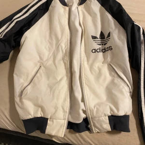 Adidas bomberjacka i svart och vitt - Snygg bomberjacka från Adidas i vitt med svarta ärmar och klassiska ränder. Jackan har dragkedja framtill, två fickor med dragkedja och stor Adidas-logga på bröstet. Perfekt för en sportig och trendig look.