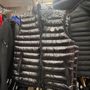 Svart dunväst från Moncler (KAN DISKUTERAS OM PRIS) - Snygg svart dunväst från Moncler med glansig finish och klassisk quiltad design. Västen har hög krage, dragkedja framtill och två fickor med dragkedja. Ikonisk Moncler-logga på bröstet och coolt serietryck på insidan. Perfekt för lager-på-lager-stil.