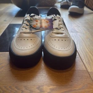 Dragon Ball sneakers från Bershka - Snygga vita sneakers från Bershka med svart sula och lila detalj vid hälen. Skorna har coolt Dragon Ball-motiv på sidan och mönstrad innersula med drakbollar. Tillverkade i syntetmaterial och har klassisk snörning framtill. Helt oanvända och billigare vid snabba köp.