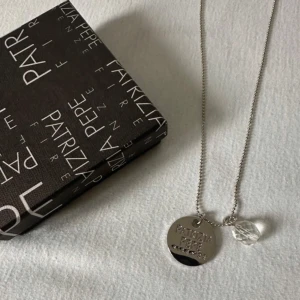 Patrizia Pepe halsband med berlock - Stilrent halsband från Patrizia Pepe med rund, blank berlock där märket är ingraverat. Kedjan är tunn och har en liten, transparent pärla som extra detalj. Perfekt accessoar för dig som gillar minimalistisk och modern stil