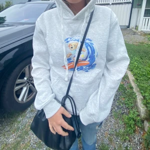Grå hoodie Polo Ralph Lauren björn - Säljer en ljusgrå hoodie från Polo Ralph Lauren med en färgglad print av en surfande björn på bröstet. Tröjan har huva med snörning, magficka och långa ärmar. Endast använd för att fota. Oroligt bra skick, skönt material.