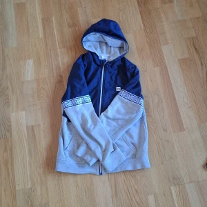 Blå och grå vindjacka från Hugo Boss - Snygg vindjacka från Hugo Boss i blått och grått med huva och dragkedja. Jackan har logoband på ärmarna och praktiska fickor framtill. Perfekt för dig som gillar sportig och stilren look. Materialmix av polyester och bomull.