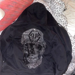 Svart hoodiejacka från Philipp Plein - Svart hoodiejacka från Philipp Plein med dragkedja, huva och snörning. Baksidan har en stor döskalle i glittriga paljetter och logga. Framsidan har diskret logomärke på bröstet. Tillverkad i mjuk bomull, perfekt för en edgy streetstil.Nypris cirka 15000kr. Platinum cut Diamond. I fint skick har inga fläckar eller inga skador. Vi har inte andvänt den så jätte mycket men i bra skick. 