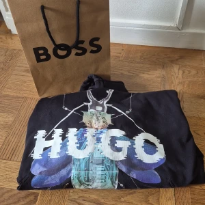 Svart HUGO BOSS hoodie med insektstryck - Svart hoodie från HUGO BOSS med stor futuristisk insekt och glitchad HUGO-logga på bröstet. Tröjan har huva med snörning och är tillverkad i mjuk bomull. Perfekt för dig som gillar streetstyle och unika prints.