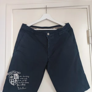 Mörkblå shorts från La Martina - Snygga mörkblå shorts från La Martina med broderade detaljer och college-inspirerat tryck på ena benet. Klassisk passform med fickor fram och bak, bälteshällor och knappstängning. Perfekta för sommardagar när du vill ha en stilren look.
