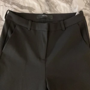 Svarta kostymbyxor från Vero Moda - Klassiska svarta kostymbyxor från Vero Moda med normal passform och raka ben. Byxorna har hällor för bälte och diskreta fickor framtill. Perfekta för en clean och stilren look. Materialet känns mjukt och följsamt.