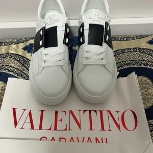 Valentino Garavani Open sneakers vit/svart - Snygga Valentino Garavani Open sneakers i vitt skinn med bred svart läderdetalj över vristen och ikoniska nitar baktill på sulan. Klassisk rund tå, platt sula och vita snören. Perfekta för dig som vill ha en stilren men ändå lyxig look.